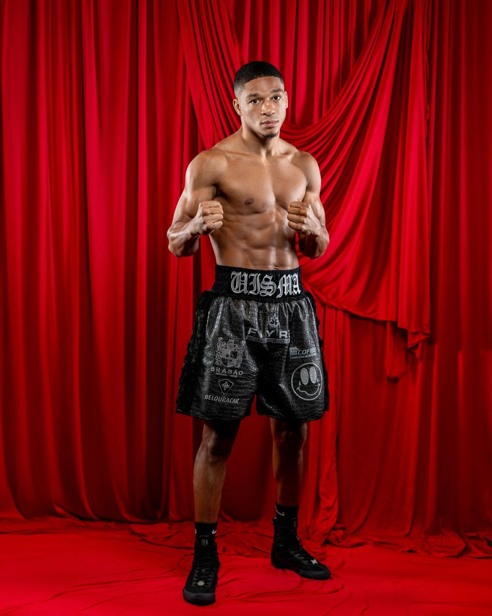 Shirtless boxer in black shorts poses in front of red curtain backdrop.