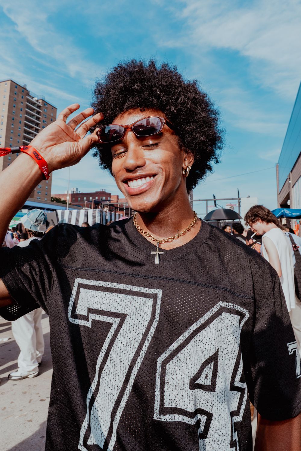 A stylish man with an afro adjusts his sunglasses wearing a black number 74 jersey at an outdoor event.