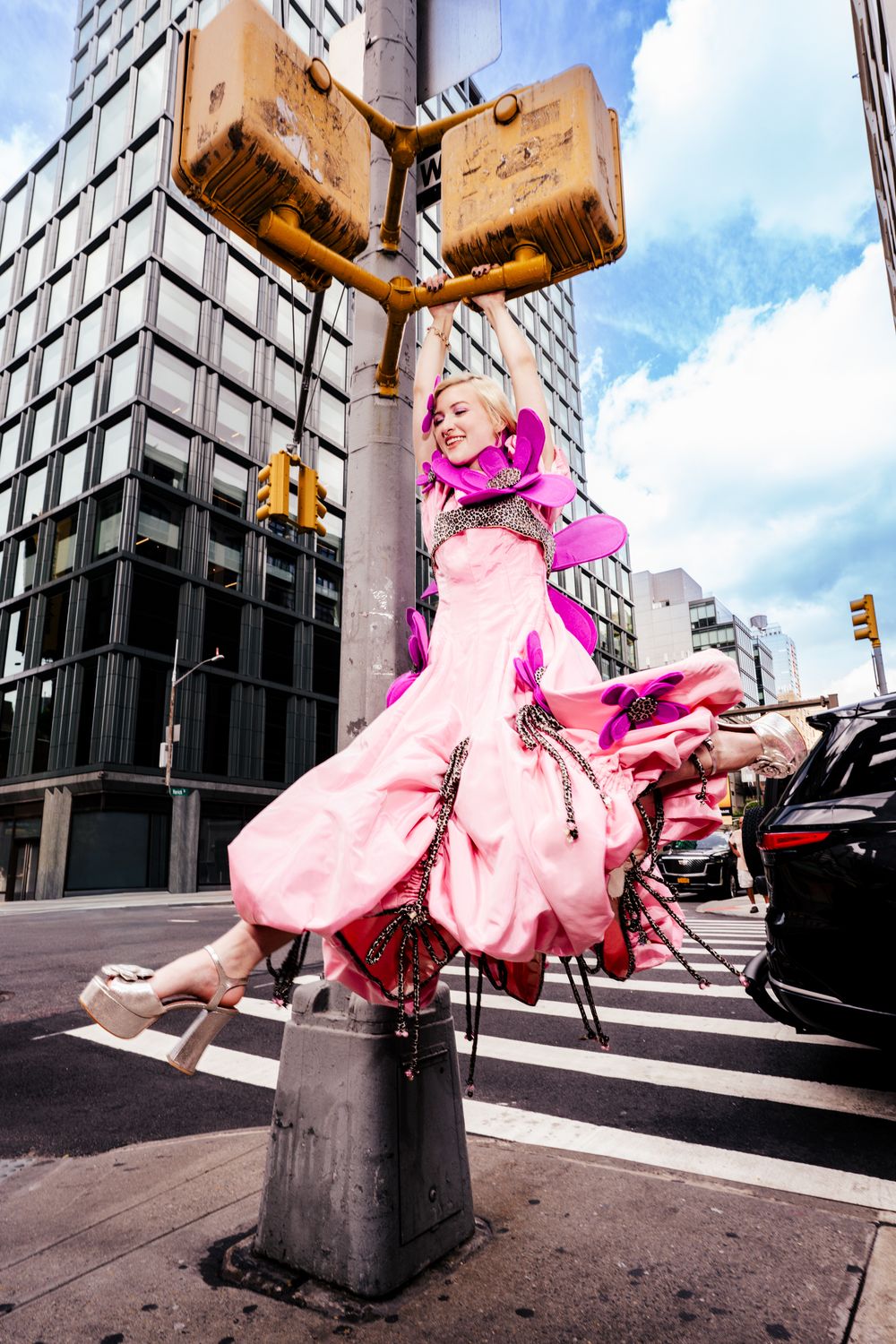 Woman in pink gown hangs from traffic light pole on city street.