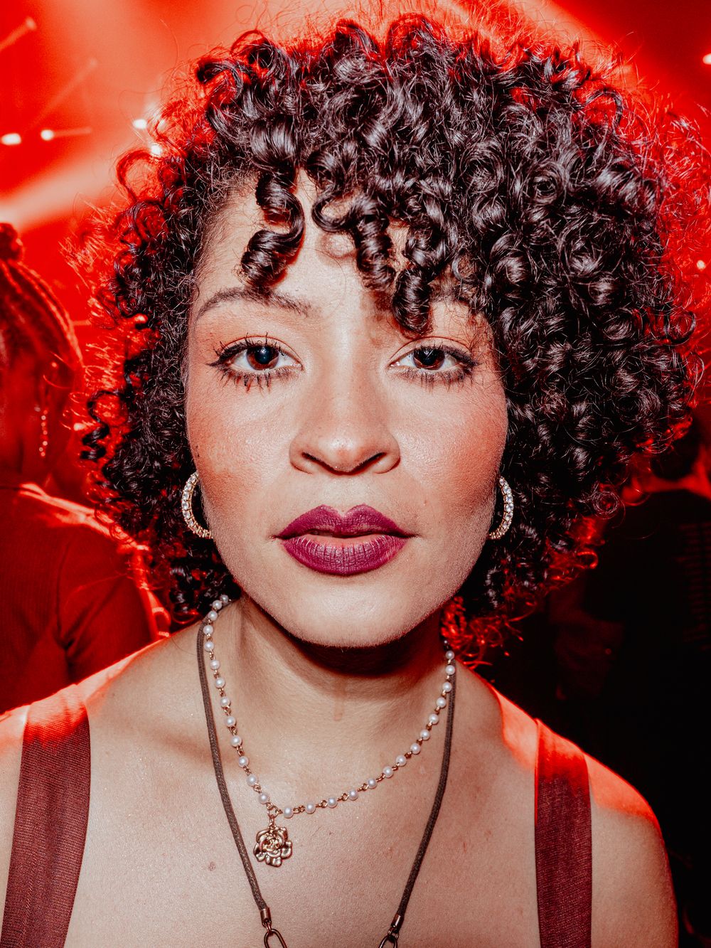 Woman with curly hair and purple lips wearing hoop earrings in red light.