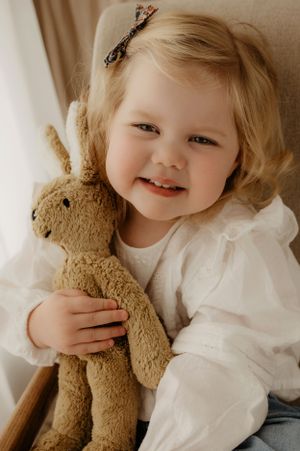 A child in a white dress holds a beloved brown plush bunny toy while sitting on a beige chair.