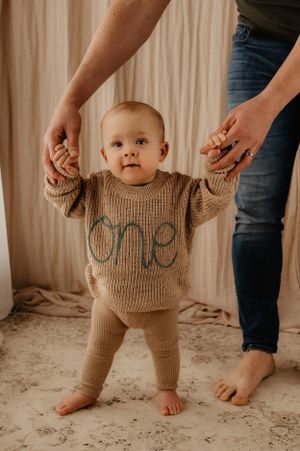 Baby in beige knit outfit stands holding parent's hands against curtained backdrop.