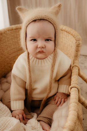 Baby wearing cream knit sweater and fuzzy bear hat sitting in wicker chair.