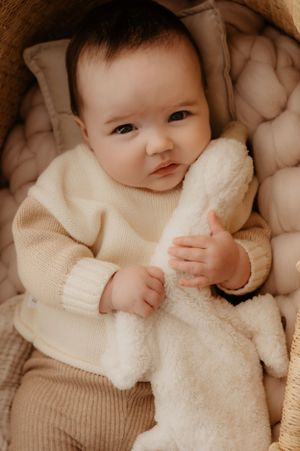 Baby in cream sweater holding soft blanket while lying in plush chair.