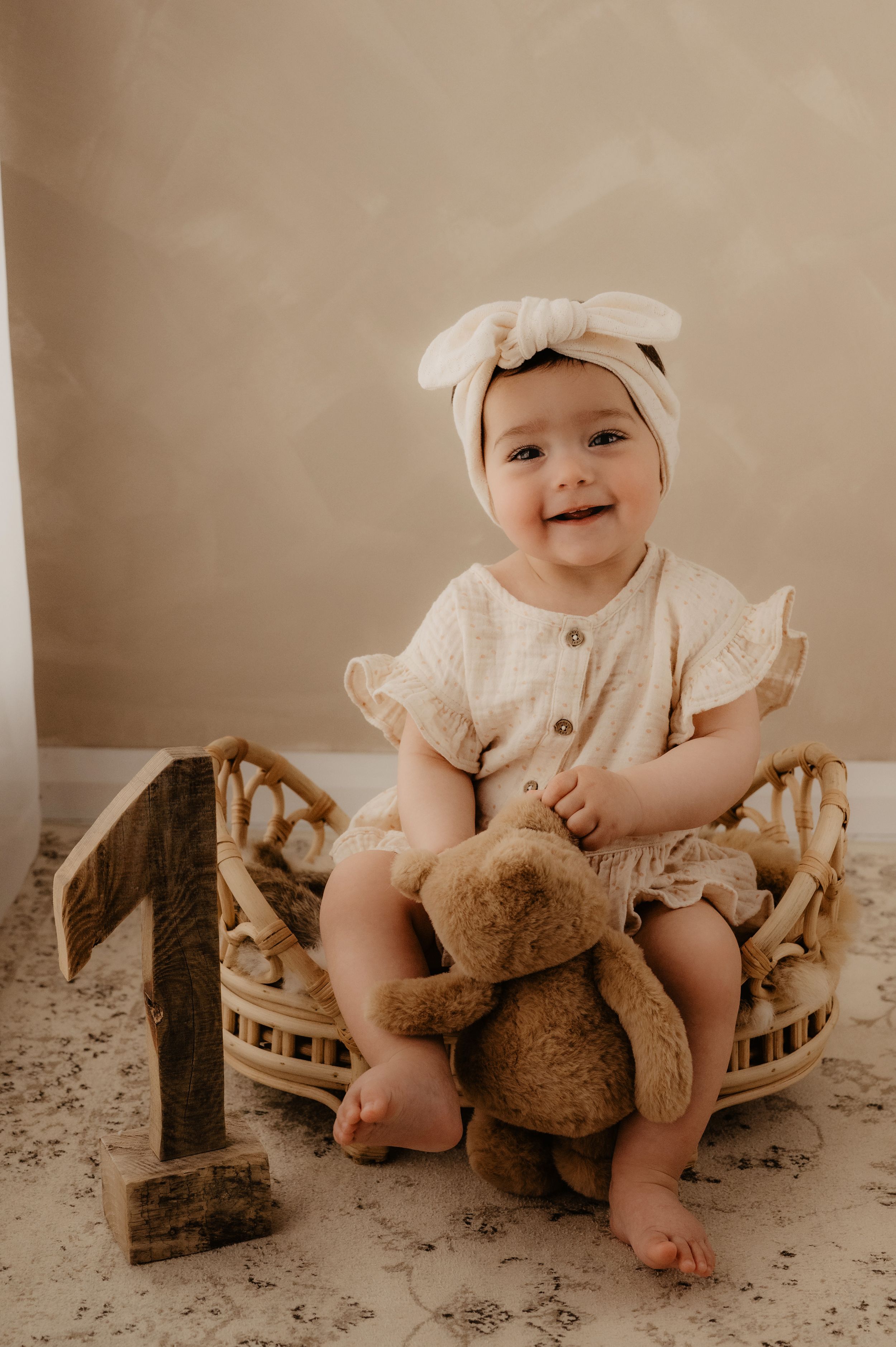 Baby in white dress plays with teddy bear on patterned floor mat.