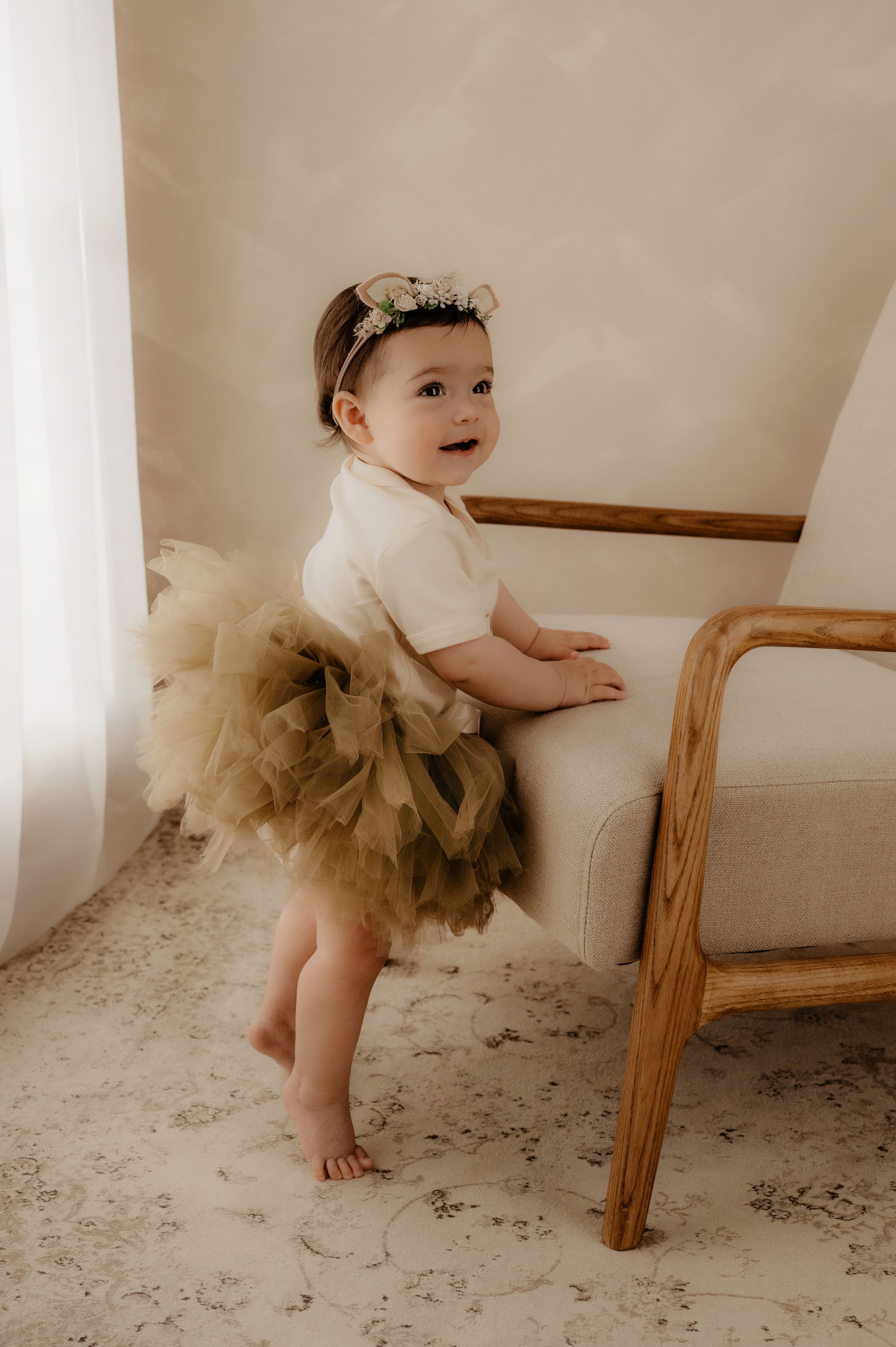 Portrait of a child in a white top and brown tulle skirt leaning against wooden furniture.