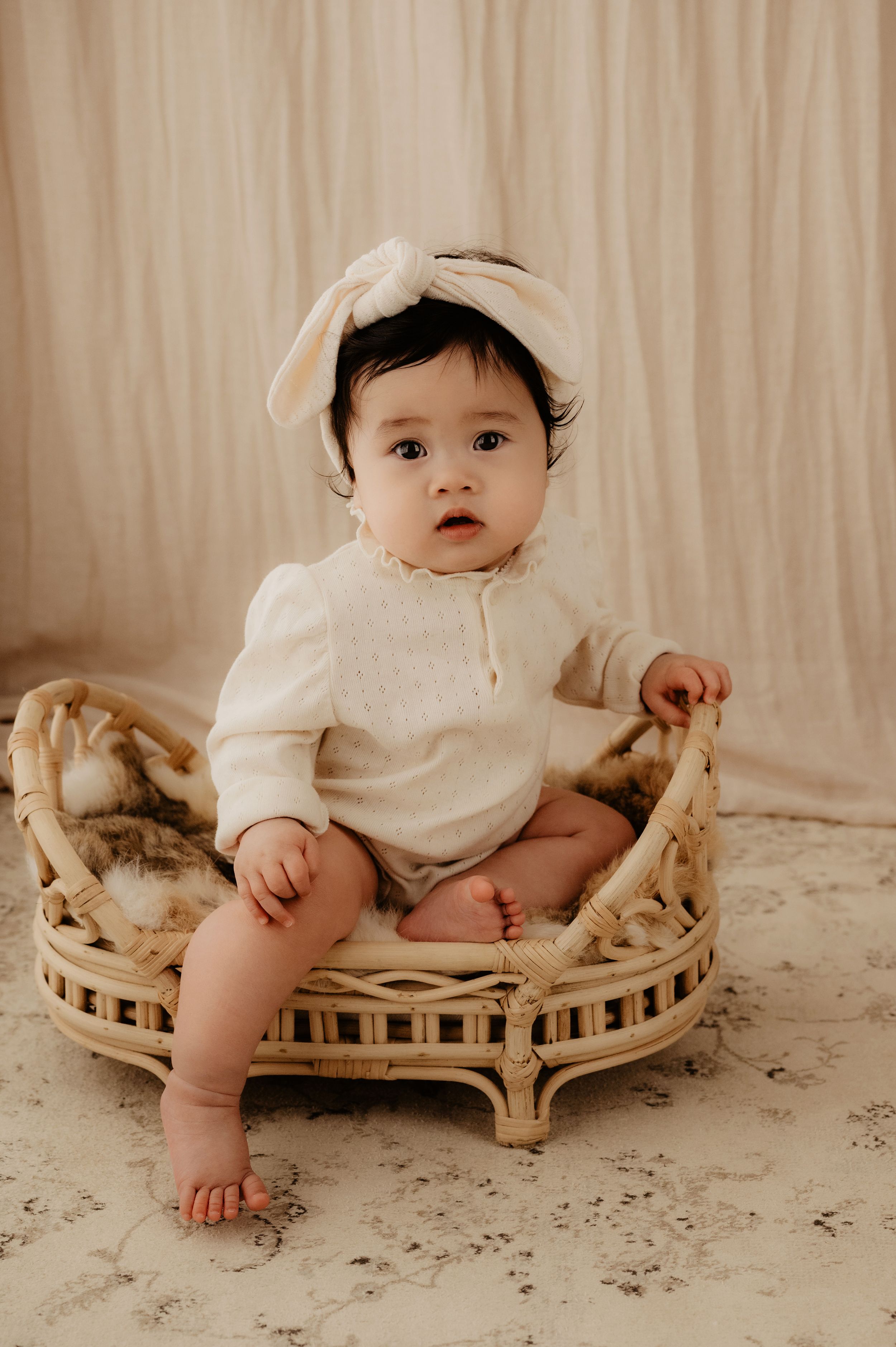 Small baby wearing white romper and bow poses in rattan chair against neutral background.
