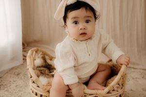 Baby in white outfit and bow sitting in vintage wicker chair during portrait session.