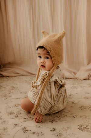 Baby in beige knit bear hat and ruffled outfit sits on patterned floor during photo session.