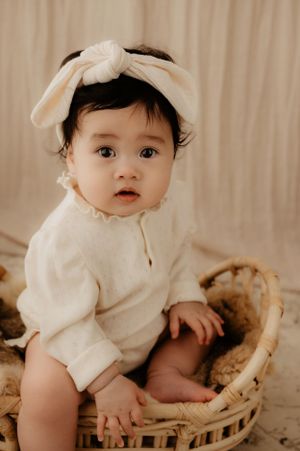 Baby wearing cream outfit and bow headband sits in wicker basket against beige backdrop during portrait session.