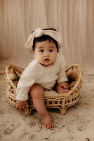 Baby in white outfit and bow sits in wicker chair during professional photoshoot.