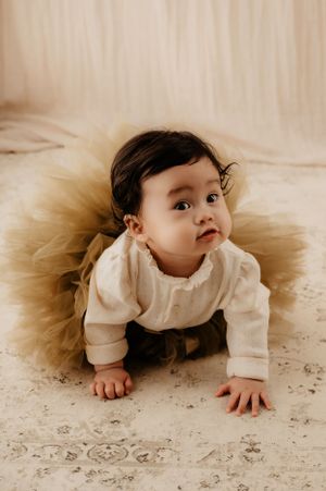 Baby in cream outfit with brown tulle skirt crawls on patterned floor during photoshoot.
