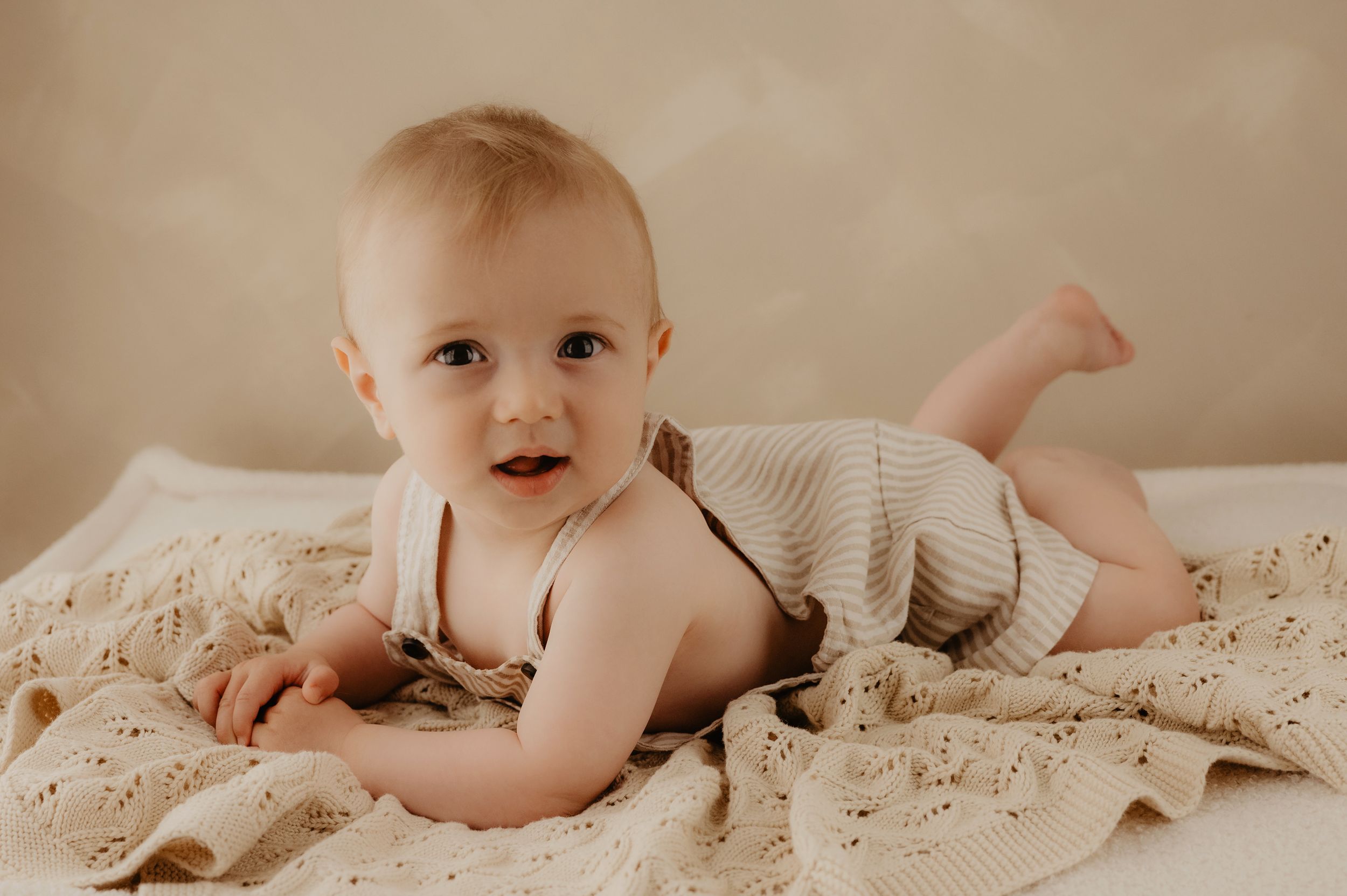 A baby lying on a beige blanket wearing a knit romper in a warm-toned portrait photo.