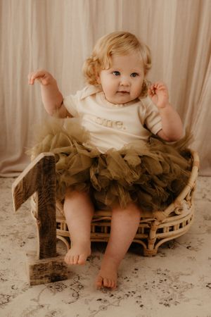 Baby in white outfit with brown tutu sitting on decorative cake smash set against cream backdrop.