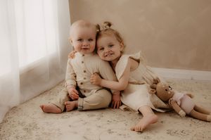 Two young children in light colored outfits sit together on a beige carpet with a teddy bear.