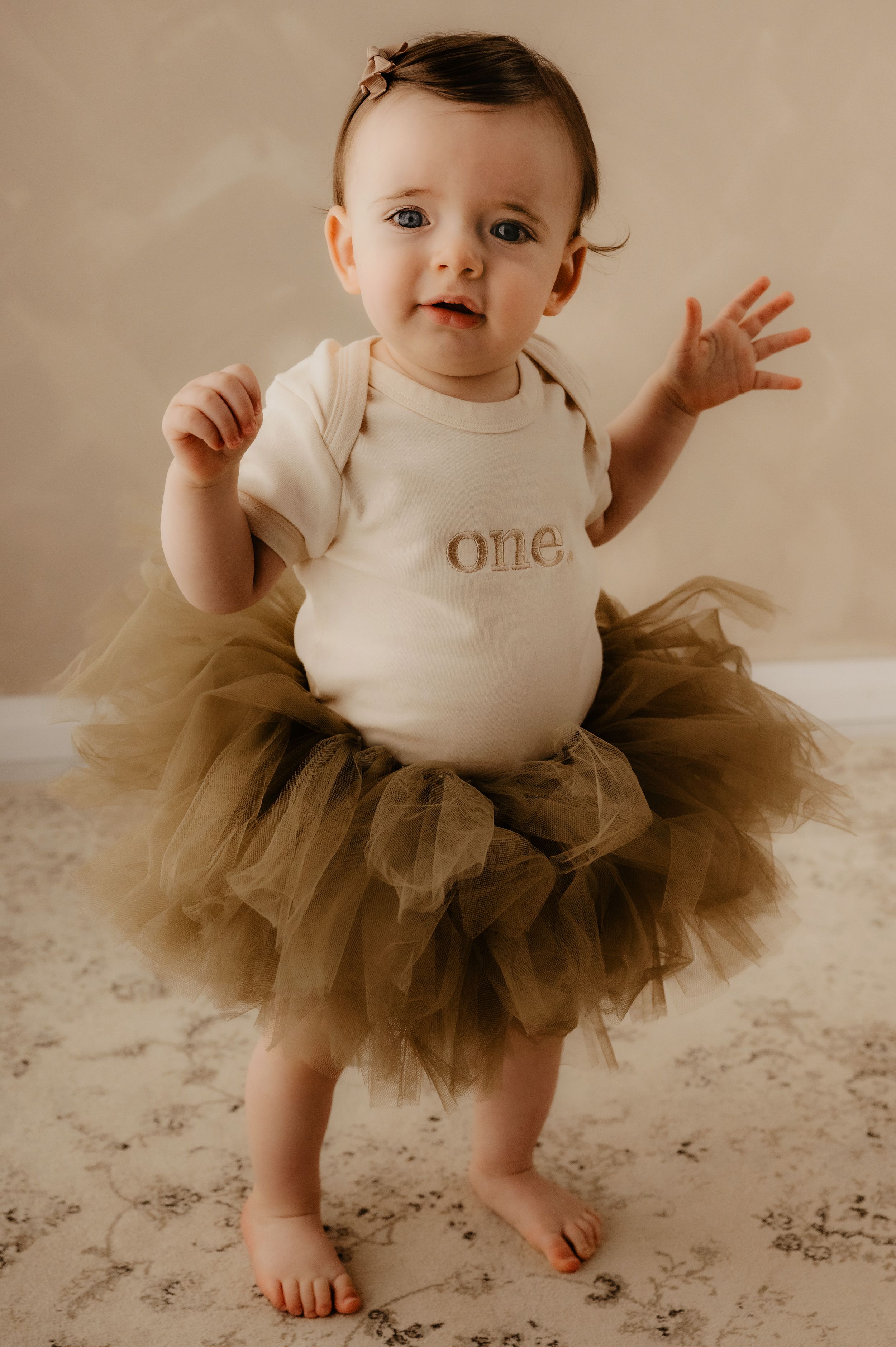 Baby wearing white onesie and brown tulle skirt stands during milestone photo shoot.