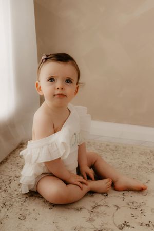 Baby in white ruffled outfit sits on textured floor during portrait session.