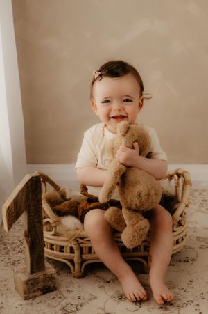 Smiling baby sits in woven basket holding teddy bear during photo session.