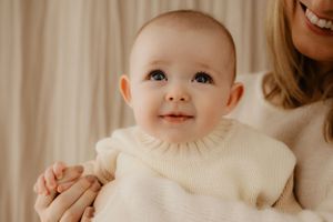 Baby wearing cream sweater smiles brightly against beige background.