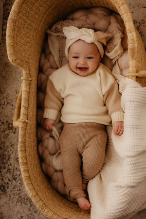 Baby in cream sweater and brown pants lies smiling in woven basket with cozy blankets.