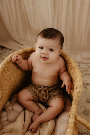 Happy baby sitting in a cozy woven basket wearing brown knit shorts against a beige backdrop.