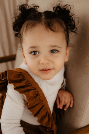 Baby wearing brown ruffled outfit and white turtleneck smiles at camera.