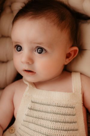 Close up portrait of baby in tan knit romper against neutral toned cushioned background.