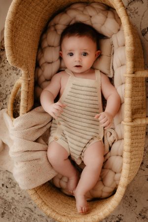 Baby wearing striped romper rests in cozy woven bassinet with quilted padding.