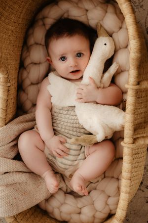 Baby in knit romper cuddles white plush duck toy in woven basket with quilted cushioning.