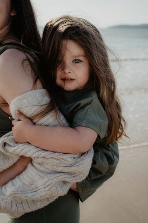 A young child in a green shirt hugs a cream-colored blanket while standing on a beach at sunset.