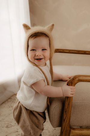 Smiling baby in bear bonnet and casual wear holding onto furniture in bright setting.