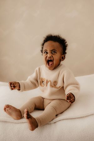 Baby wearing cream ribbed one-piece outfit laughs joyfully while lying on white bedding.