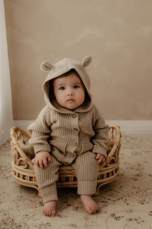 Baby wearing beige knit hooded outfit sits in woven basket during professional photoshoot.