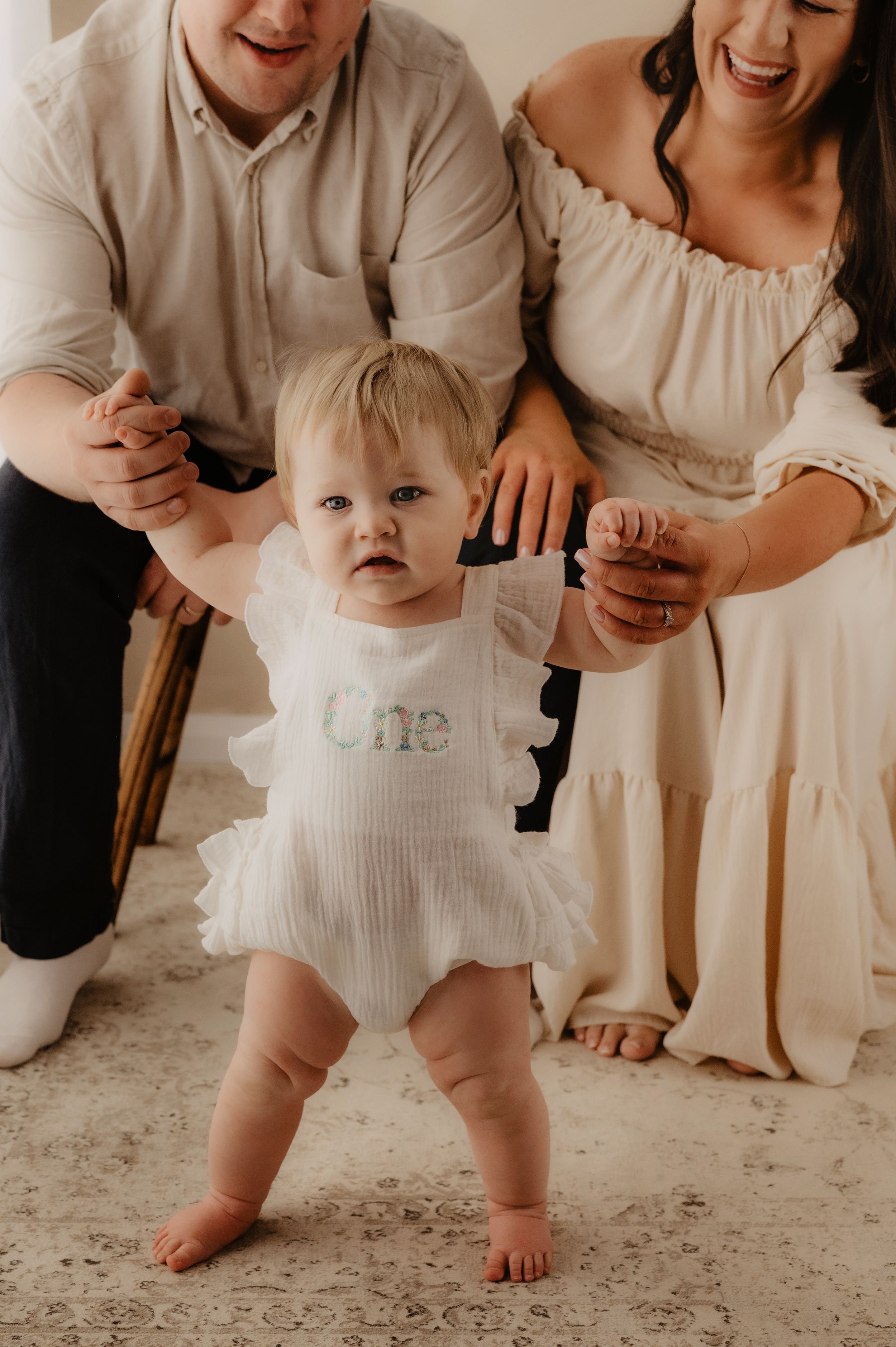 Toddler in white lace dress takes first steps with parents holding hands in rustic indoor setting.