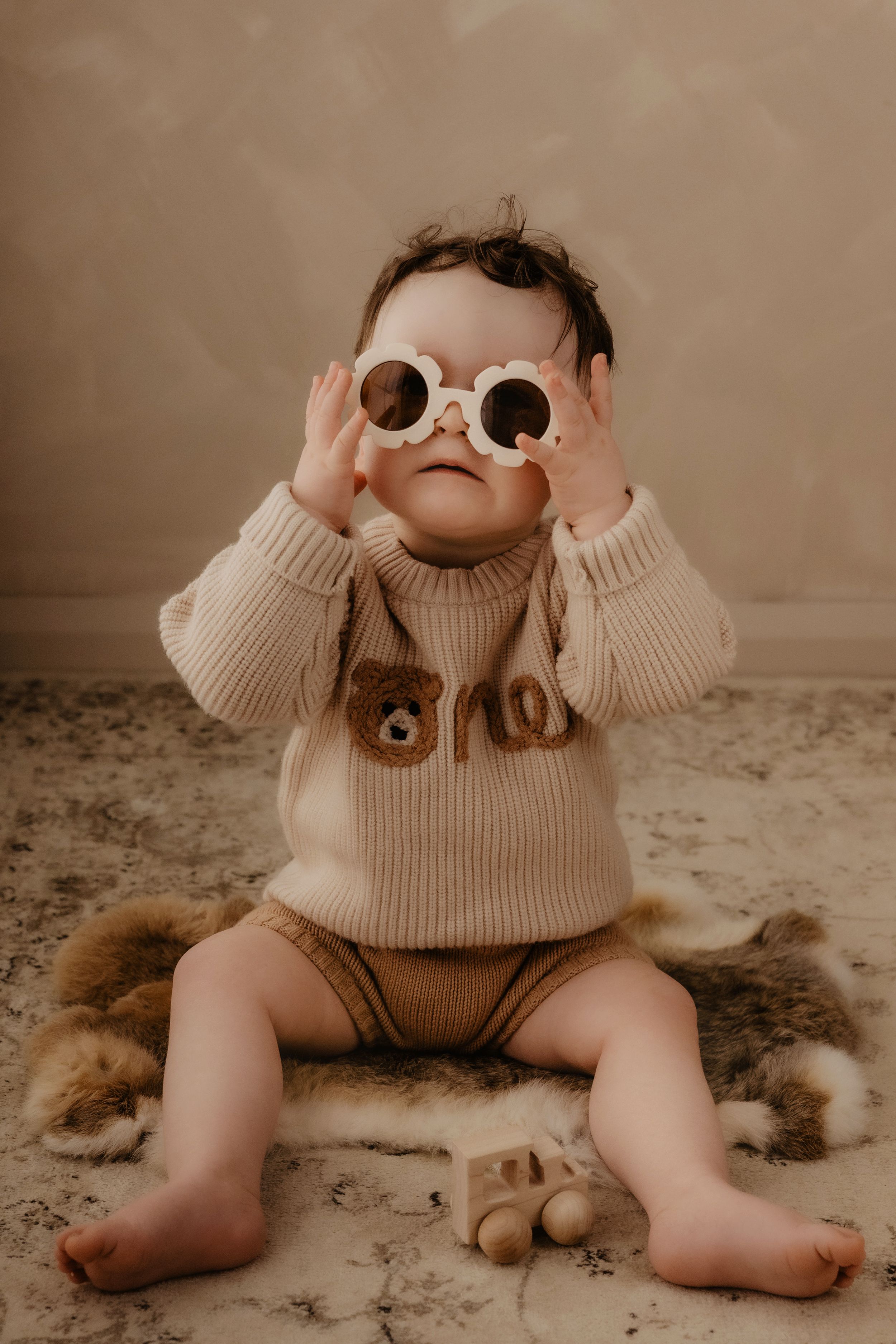 Baby in cream sweater plays with sunglasses while sitting on floral rug in artistic photo series.