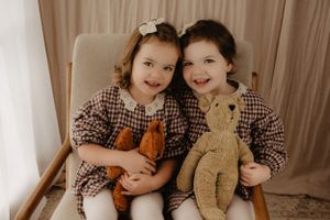 Two children in matching gingham dresses sitting together with stuffed animals on a beige chair.