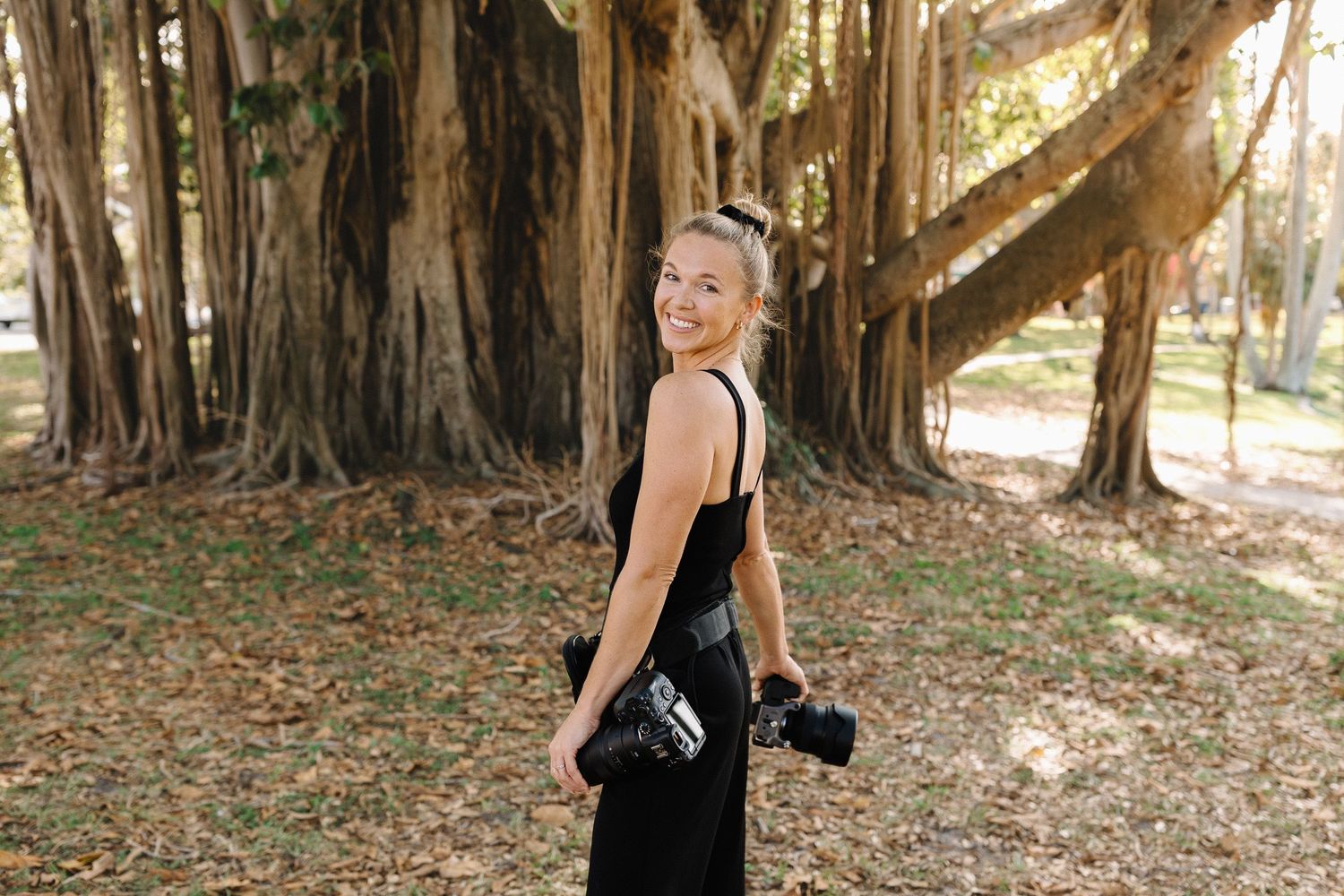 A smiling female photographer in a black dress holds her camera while posing outdoors near large banyan trees.