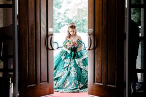 XVin stunning emerald green ball gown with lace details holds colorful bouquet standing in ornate wooden doorway. Charleston, Summerville,So
