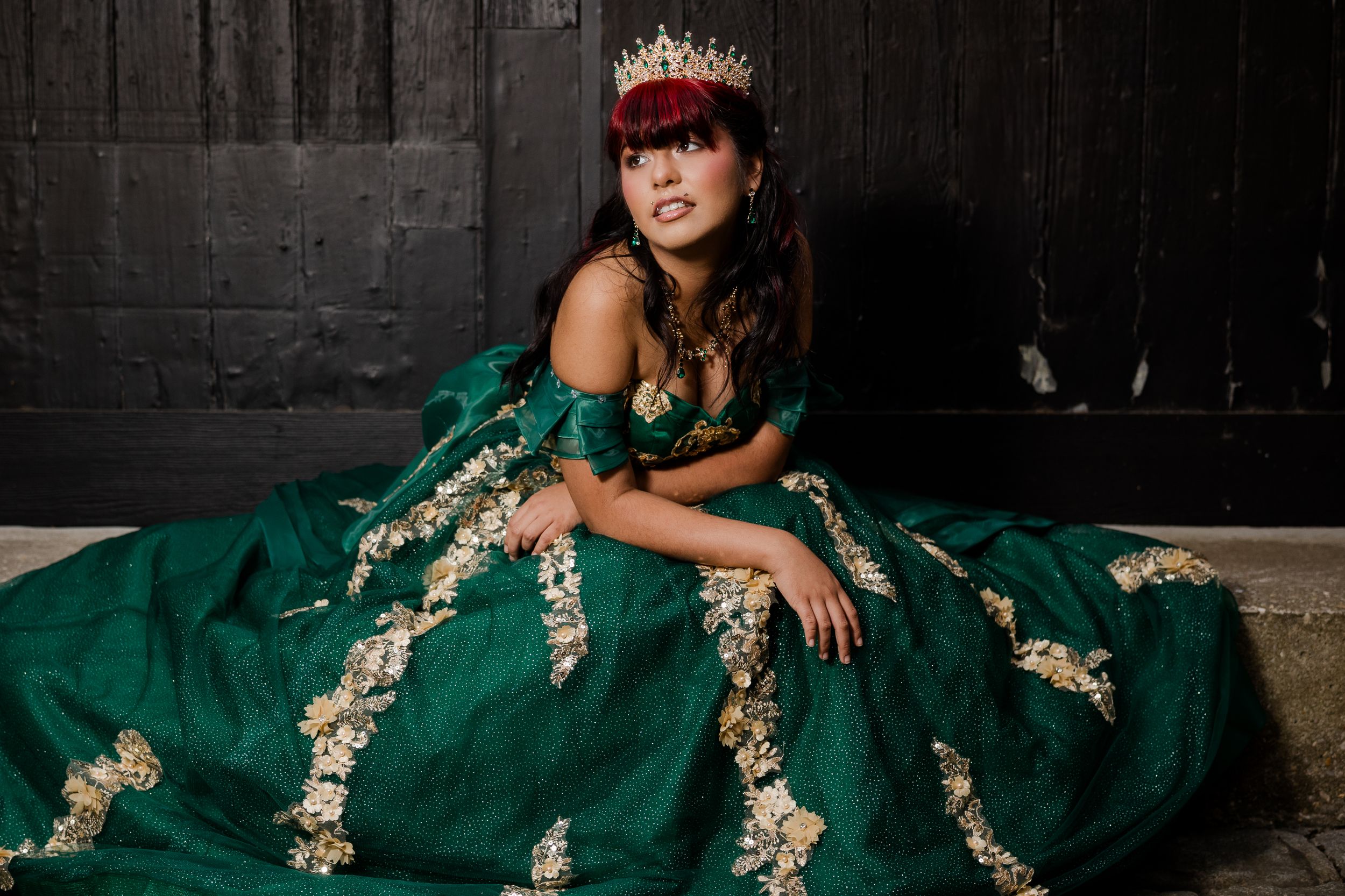 Young woman in elegant emerald green quinceañera ball gown with gold embroidery and red floral crown poses gracefully. Charleston, SC