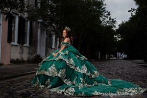 Young woman in emerald green quinceañera ball gown with gold embroidery and tiara poses on cobblestone street. Charleston, Summerville SC