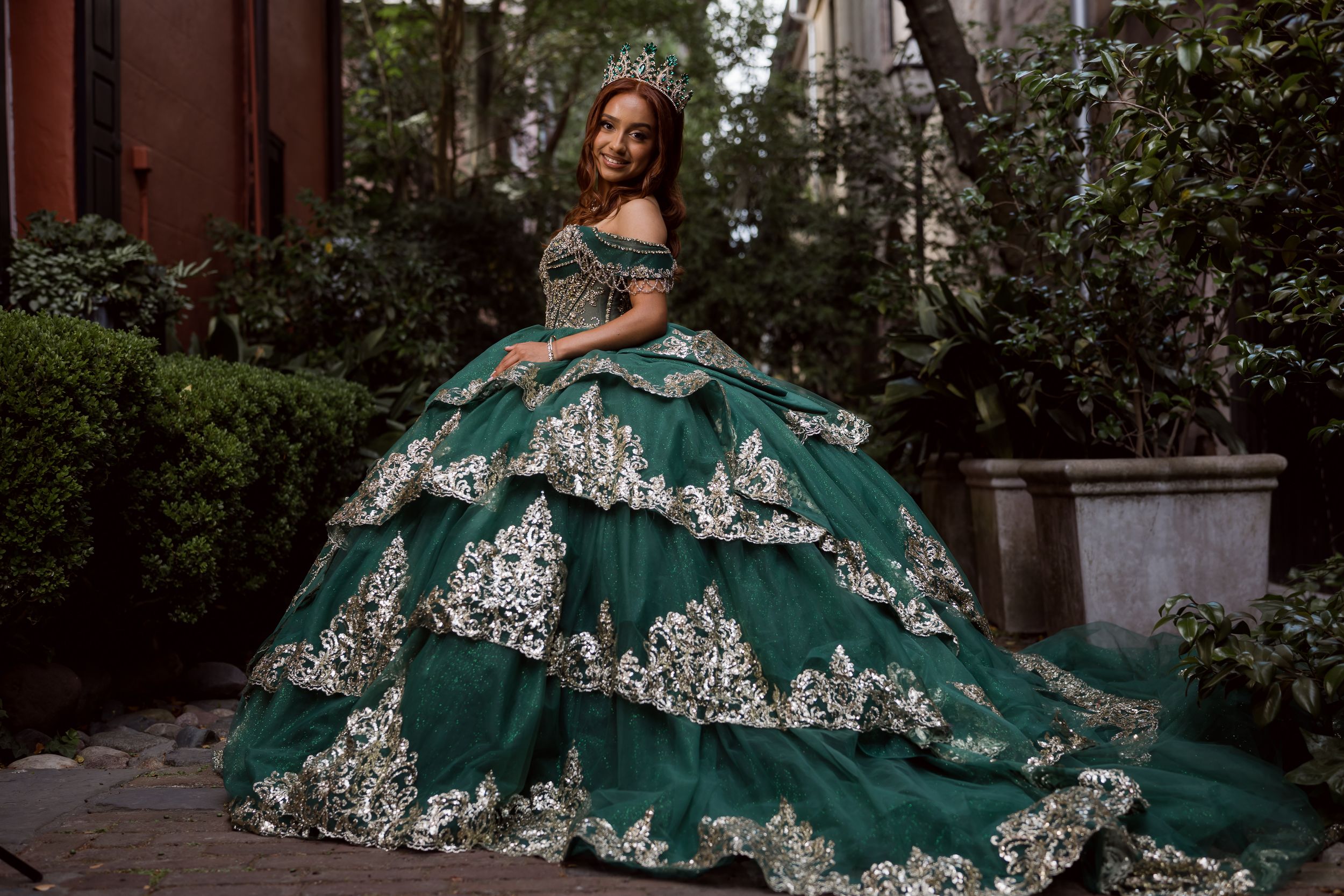 Woman in emerald green ball gown with gold lace appliques and off-shoulder neckline poses in garden setting. Charleston, Summerville, SC