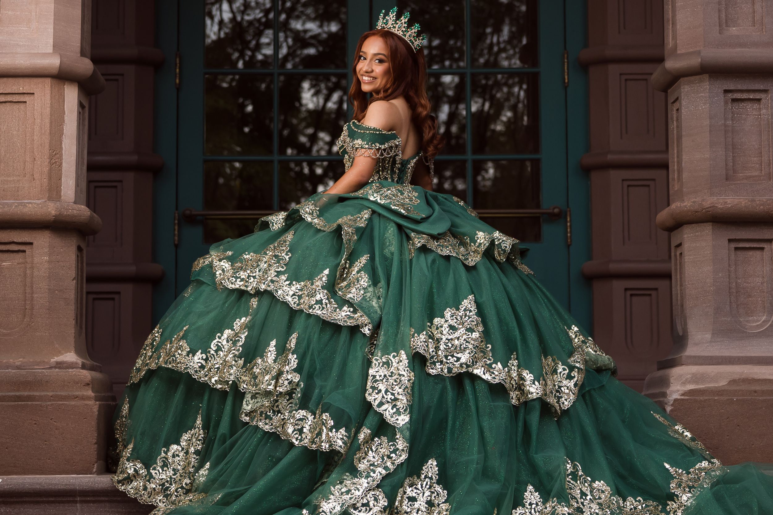 Woman in emerald green ball gown with gold lace appliques and tiered skirt, wearing a crown, posing outdoors. Charleston, South Carolina