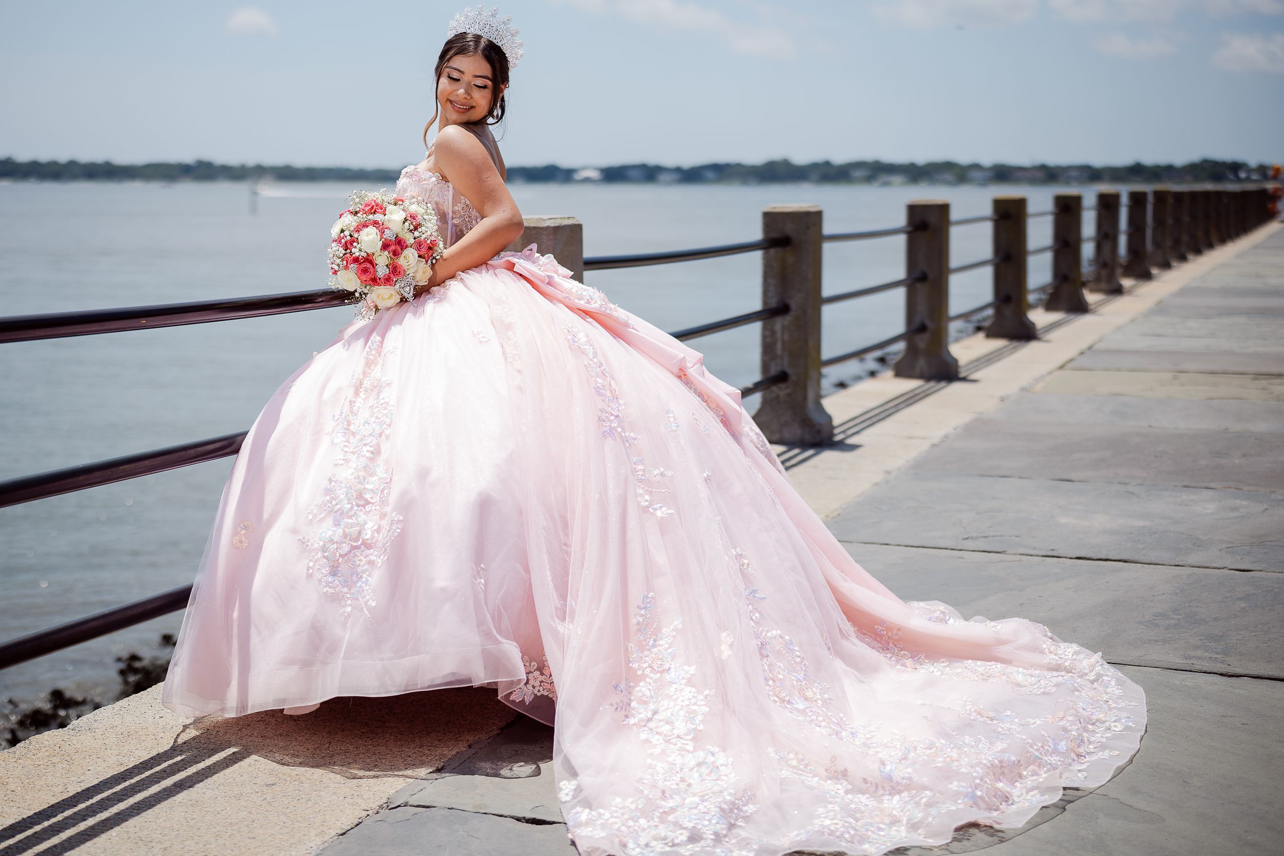 A quinceanera in an elegant pink ball gown with a train poses at sunset along a waterfront railing.
