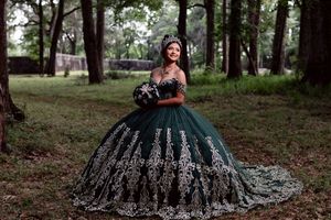 A quinceañera with a dark, elegant gown with flowing tulle fabric poses gracefully in a wooded outdoor setting.