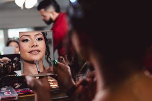 A reflection appears in a makeup mirror during a beauty session with cosmetics and brushes visible in the background.