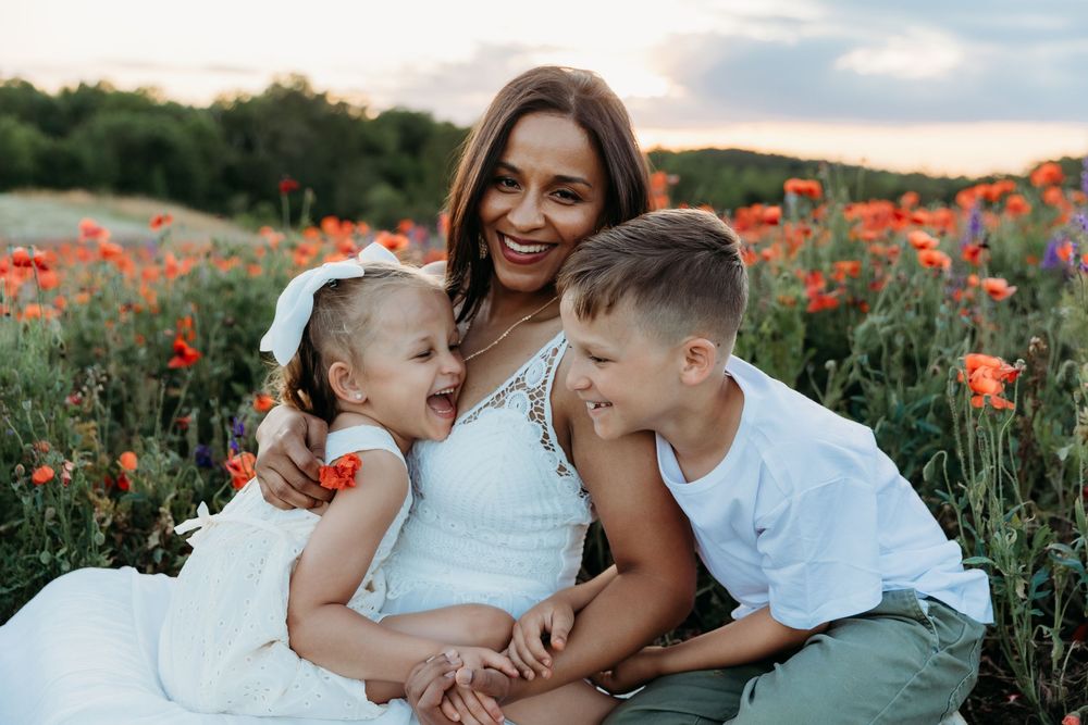 A smiling mother sits with her two young children in a vibrant poppy field at sunset.