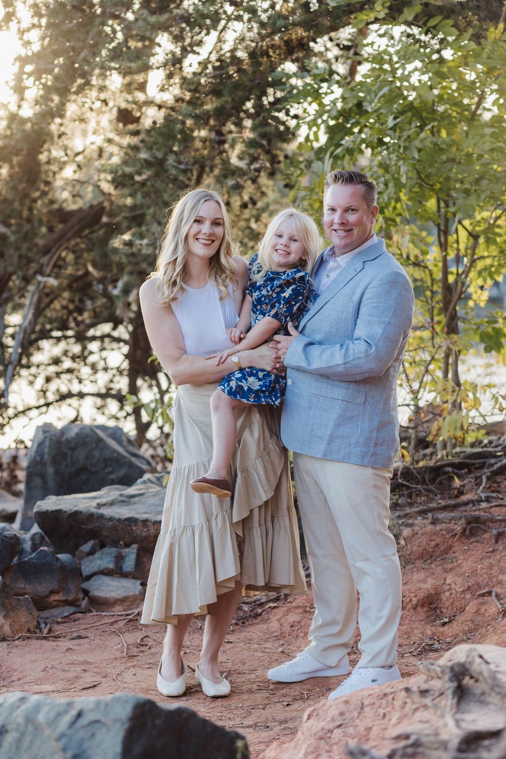 Happy family of three posing outdoors among rocks and trees in coordinated neutral and blue outfits.