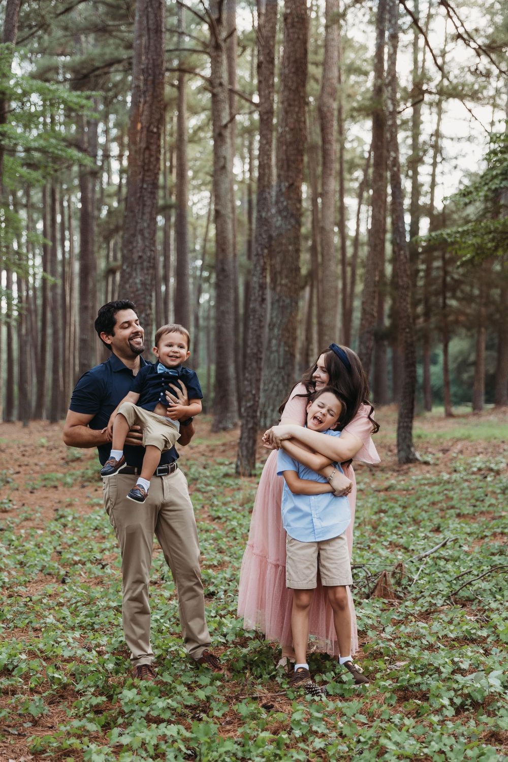 Family of four enjoying a outdoor photo session in a pine forest, parents holding children while smiling.