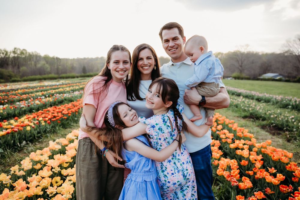 Family of six smiling together in a colorful tulip field at sunset, surrounded by orange and yellow blooms.