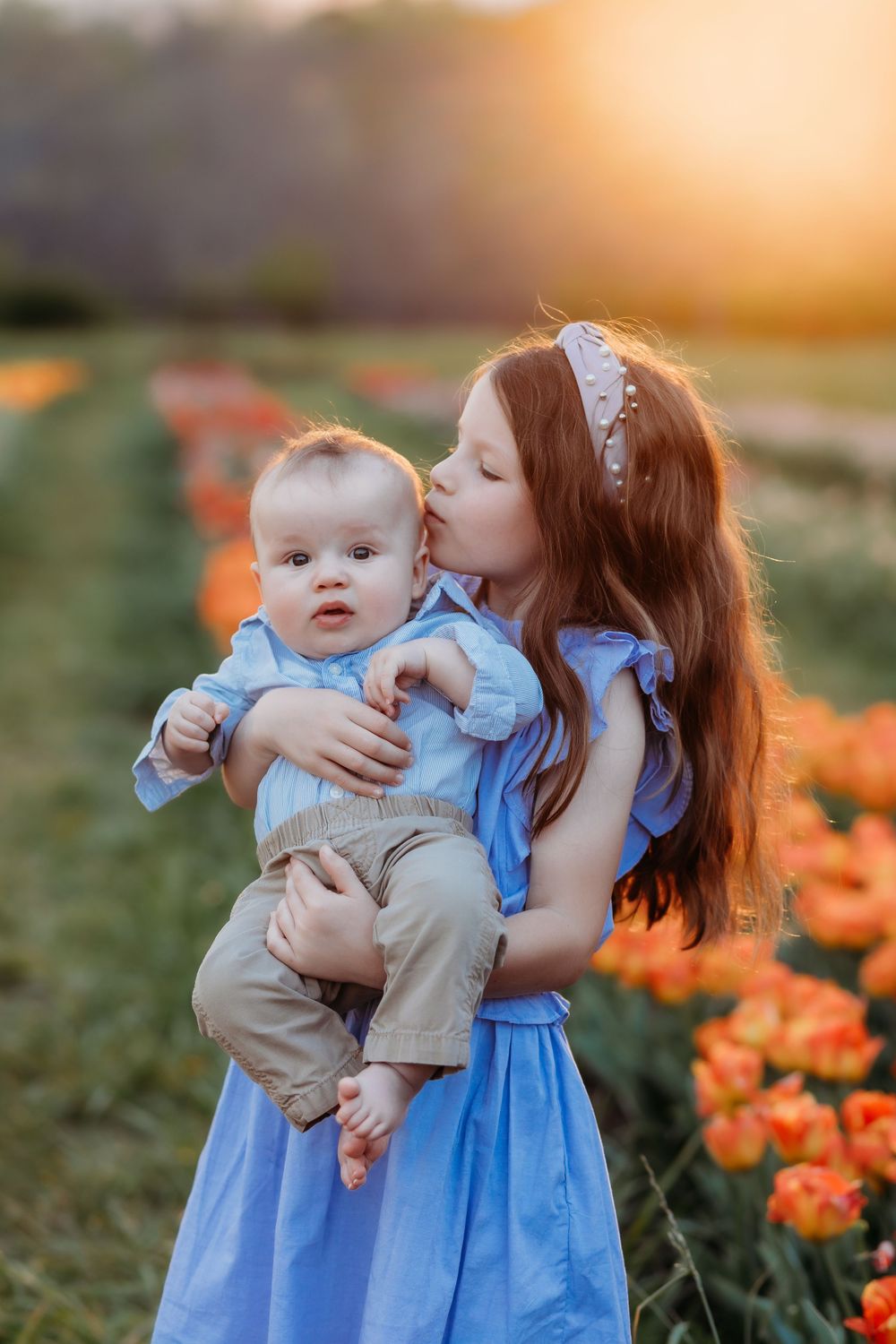 A young girl in a blue dress kisses a baby boy in a tulip field at golden sunset.